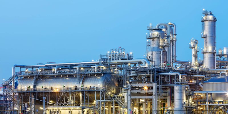 A modern, illuminated refinery is visible at dusk. Lights from the buildings and streets glow. Several distillation towers can be seen as well as glowing moon. Long exposure with tripod.